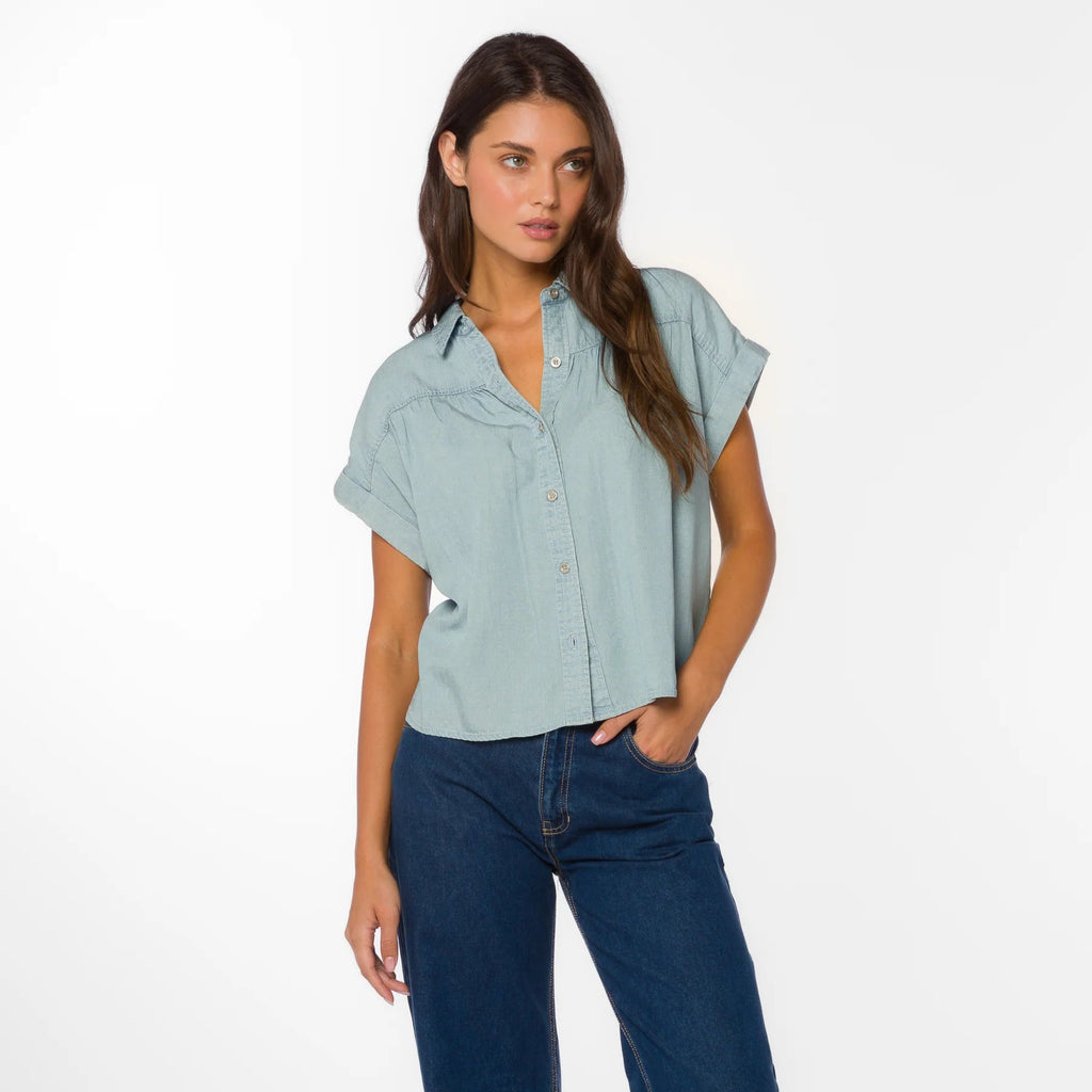 Woman wearing light blue short-sleeve button-up shirt with dark jeans, standing against white background