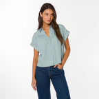Woman wearing light blue short-sleeve button-up shirt with dark jeans, standing against white background