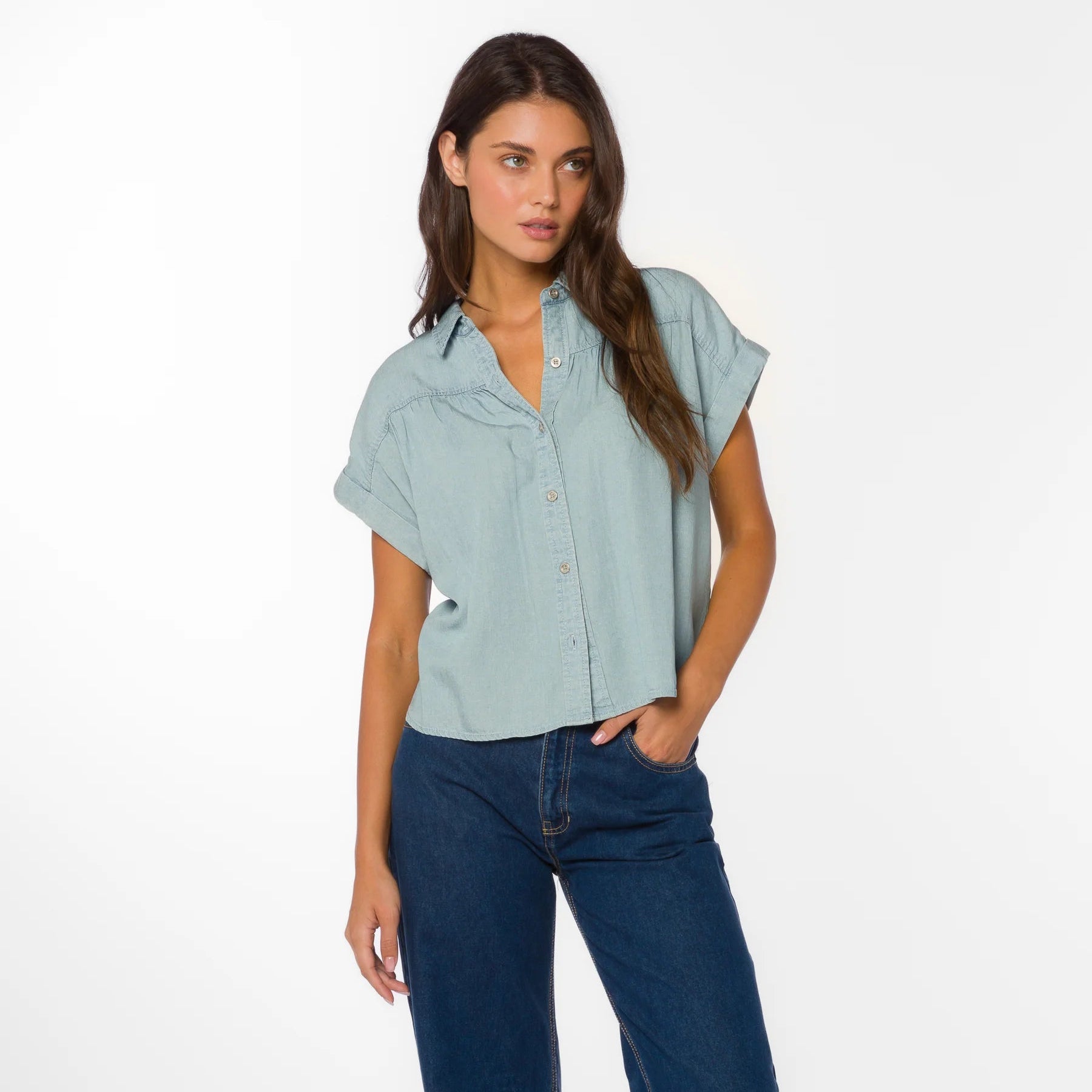 Woman wearing light blue short-sleeve button-up shirt with dark jeans, standing against white background