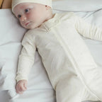 Infant wearing cream Zip Longsleeve Sleeper Footie with matching hat, lying on white bedding