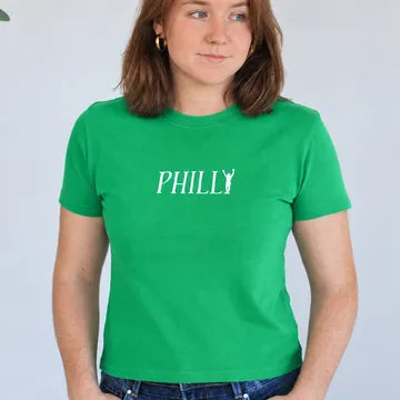 A woman wearing a bright green short-sleeve t-shirt with 'PHILLY' printed in white across the chest, paired with blue jeans, standing against a light gray wall.